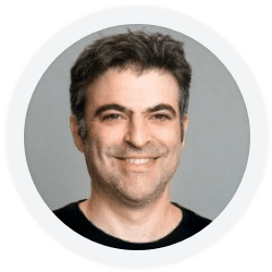 A portrait of a smiling middle-aged caucasian man with dark hair, wearing a black shirt, set against a light gray background.