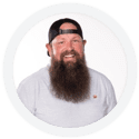 A man with a long beard and a black cap smiles while wearing a light grey T-shirt against a white background.