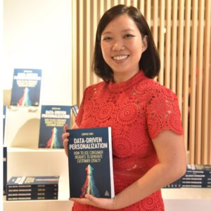 A person in a red lace dress smiles while holding a book titled "Data-Driven Personalization: How to Use Consumer Insights to Generate Customer Loyalty" against a backdrop of similar books.