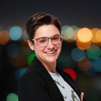 A person with short hair and glasses smiles in front of a blurred background of city lights. They are wearing a dark jacket and light-colored shirt.