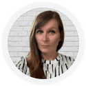 A woman with long brown hair in a white patterned shirt stands in front of a white brick wall.